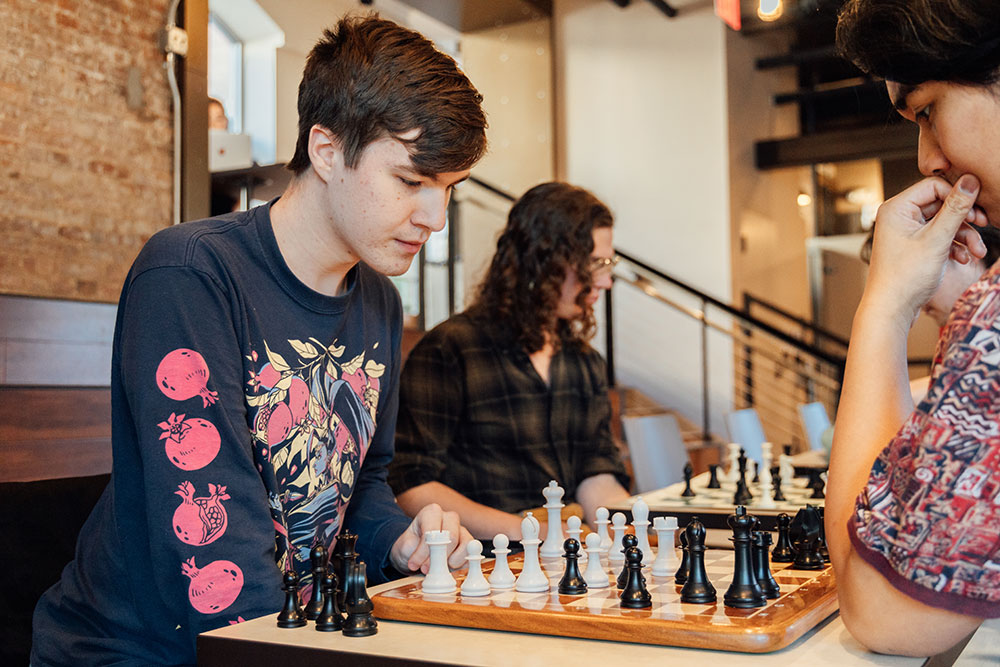 Two students play chess in the foreground while others play in the background.