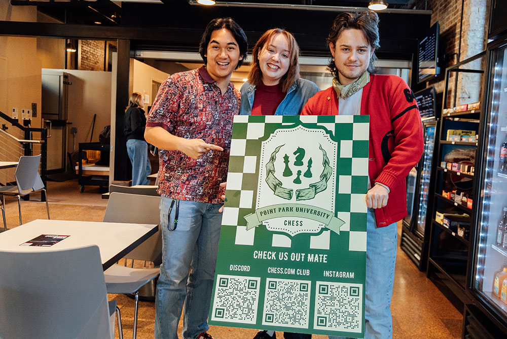 Three students stand in a coffeeshop holding a large sign advertising chess club