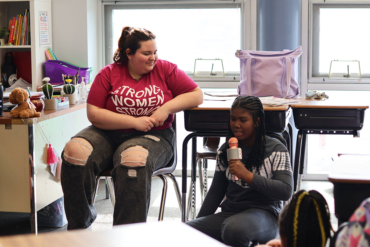 A girl speaks into a microphone while a college woman looks on.