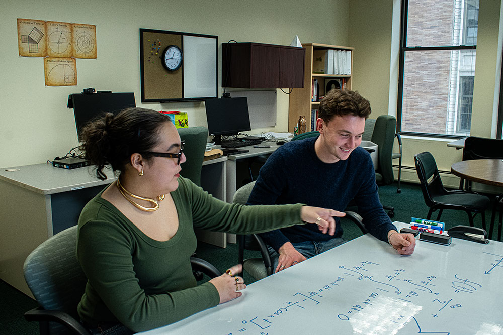 A student is tutored by a peer at a whiteboard table.