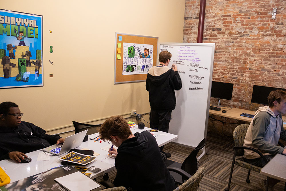A student writes on a whiteboard while others work on computers.