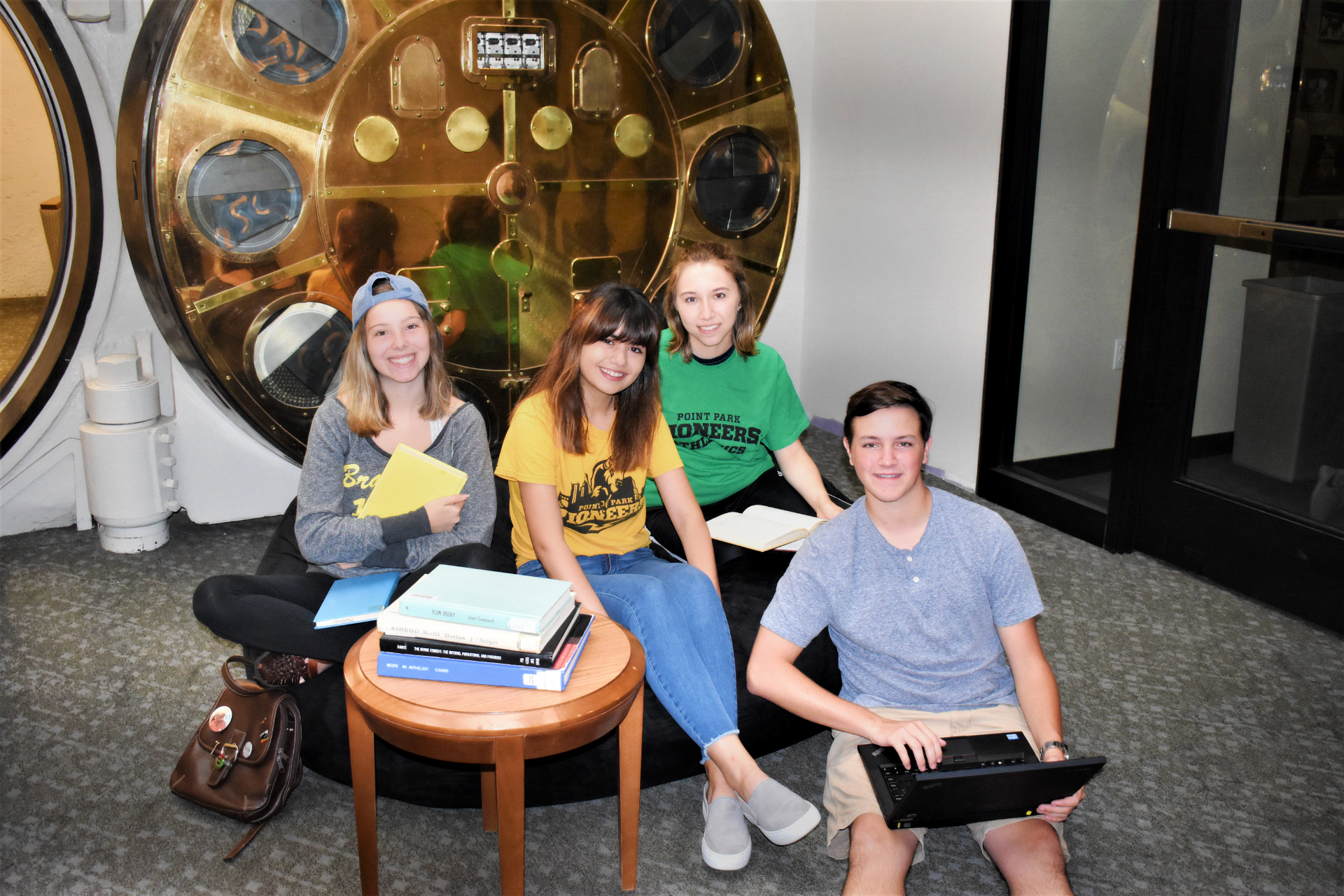 Three female and one male students sitting in the library by the old bank vault. Smiling.