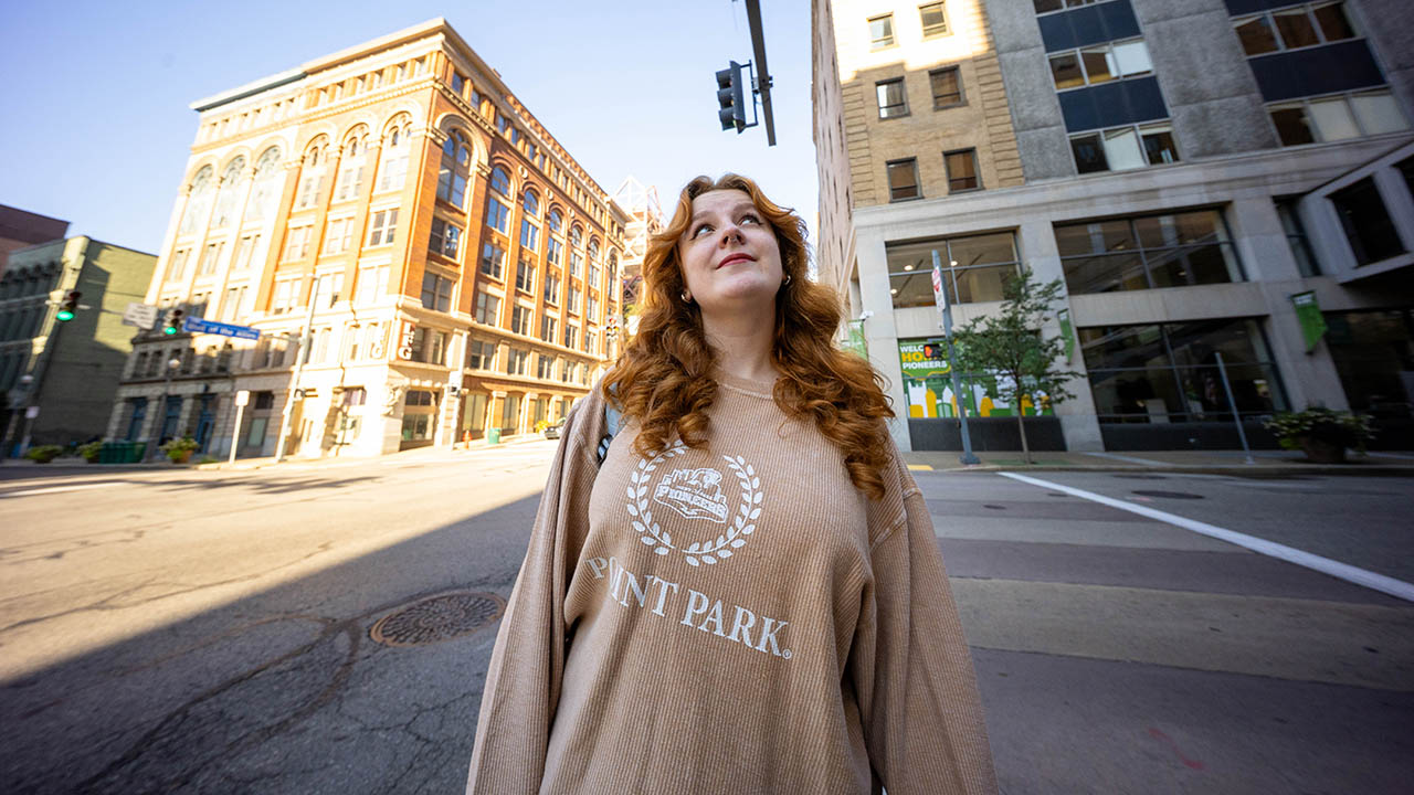Pictured is alumna Devyn Amigh in a Point Park sweatshirt looking up at a building on campus. 