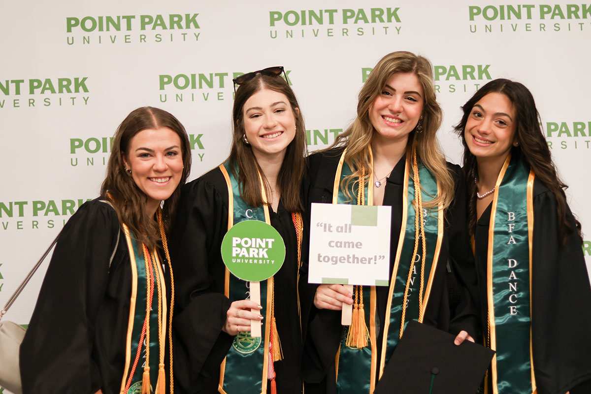 Four women in graduation gowns pose before a Point Park background