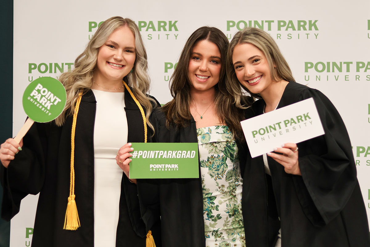 Three women in graduation gowns pose before a Point Park background