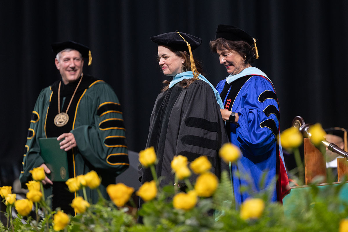 A doctoral graduate receives her hood on stage while the president looks on