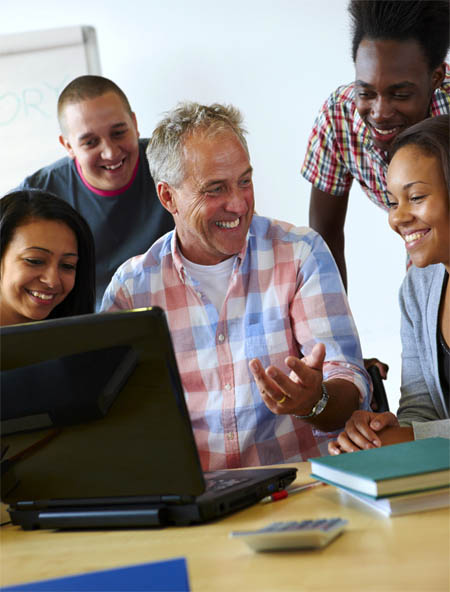 Pictured are a diverse group of students surrounding a teacher at a computer.