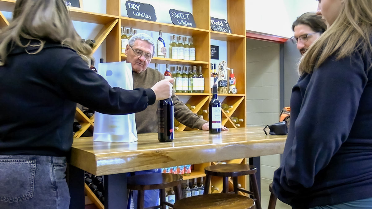 Pictured is a man behind a counter in a winetasting setting.