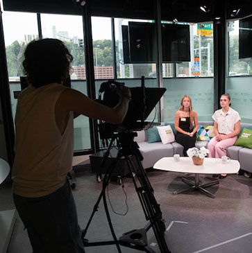 A student is behind a camera filming two female students on a newsroom set.