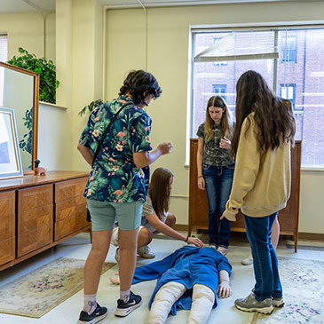 High school students stand around a dummy on the floor of the crime scene house.