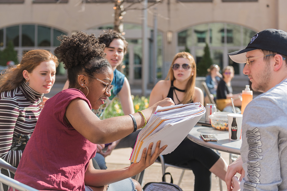 Students in Village Park gathered at a table look on as a student shares something from her notebook. 