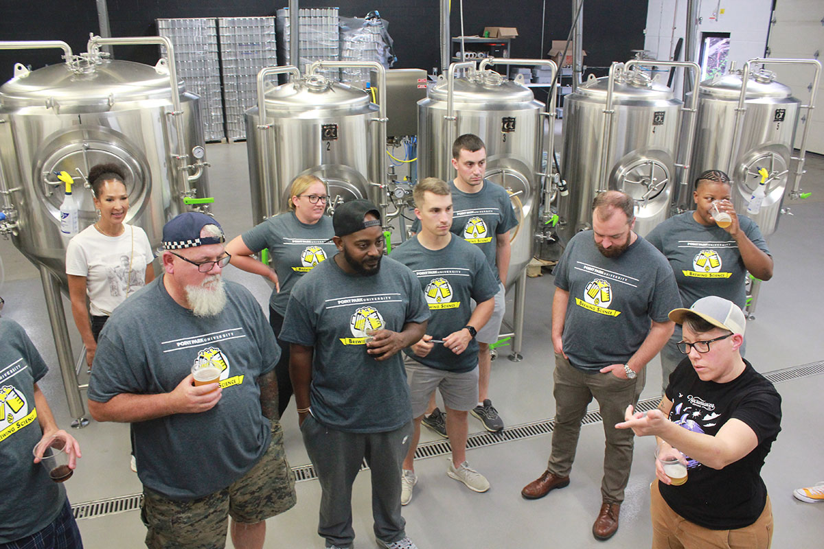 A group of people stand in a brewing room listening to instruction