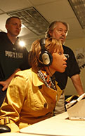 Photo of a female Point Park student seated at an audio control console during a radio production class with a Point Park faculty member offering her instruction. | Photo by Bradshaw.