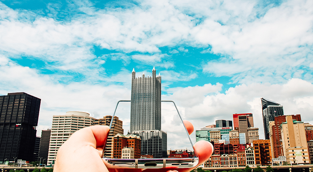 Pictured is a hand holding a cell phone toward the Pittsburgh skyline. Photo by Nathaniel Holzer. 