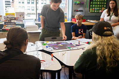A high school student demonstrates how to play his board game while other students watch.