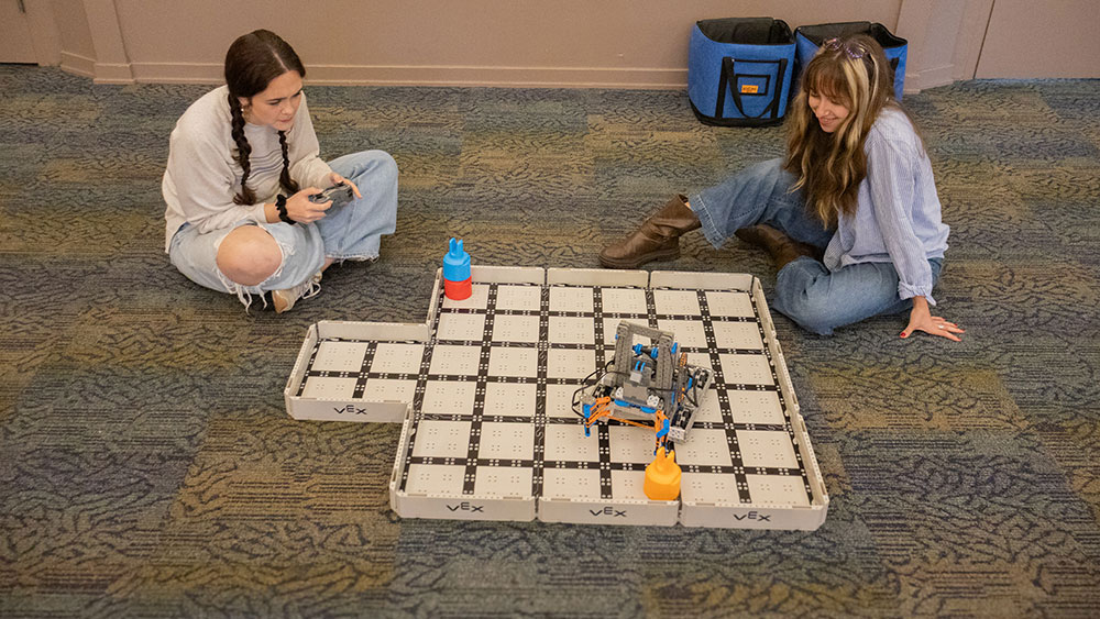 Two students sit on the floor using a remote control robot.