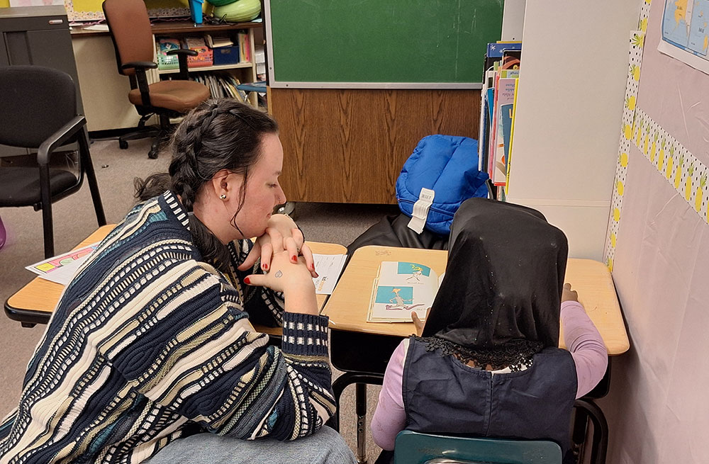 A college students works with an elementary student at a desk.