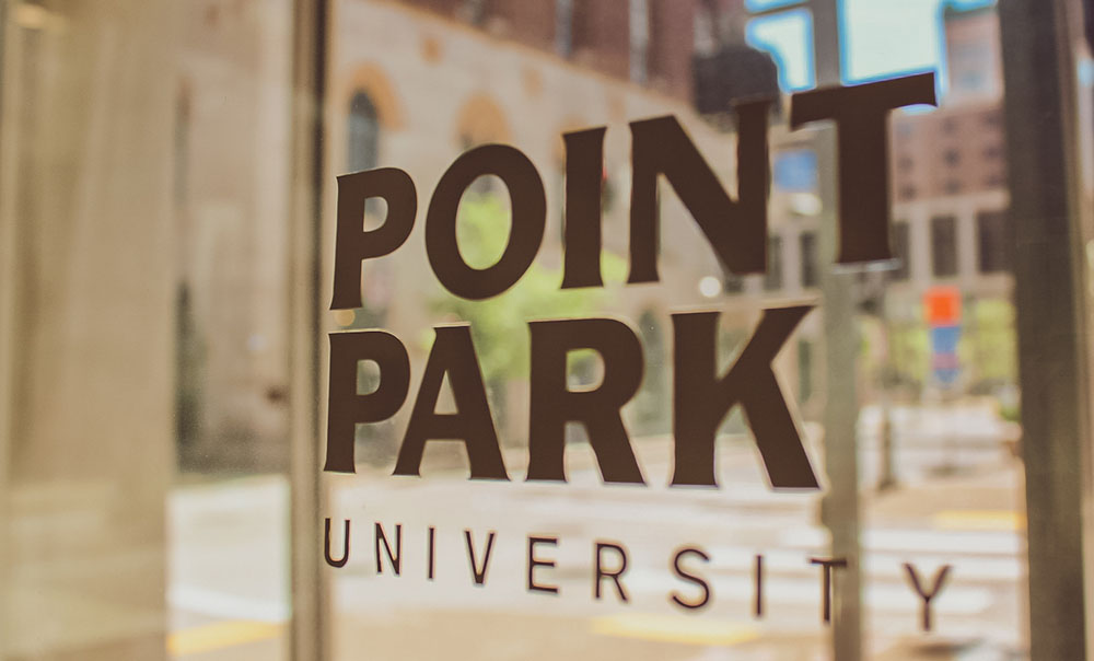 Pictured is the Point Park University logo on a glass window with the campus reflecting off of it. Photo by Nathaniel Holzer.