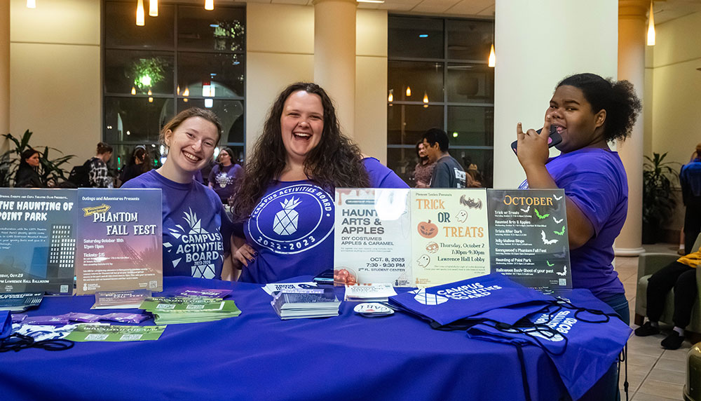 Three members of the campus activities board stand behind a table welcoming people to their event.