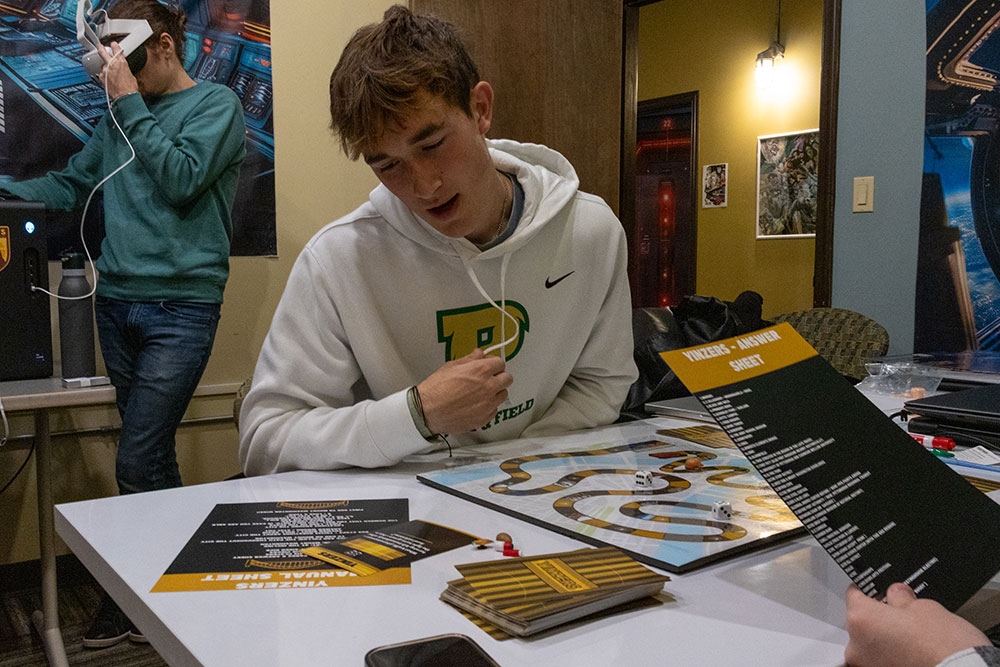 A student plays a board game while a man in the background uses VR glasses.