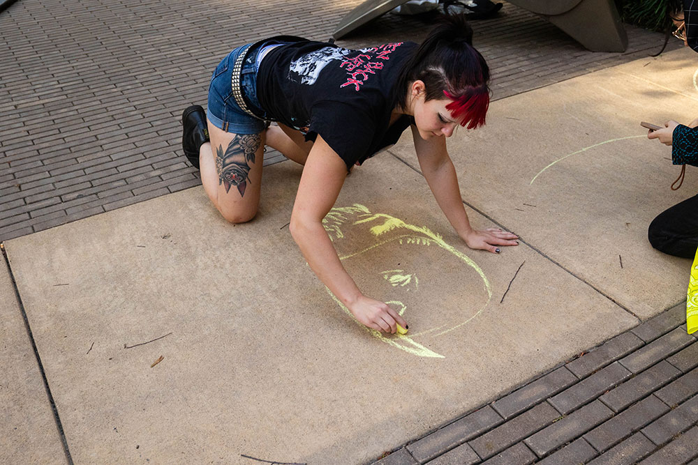A student draws a yellow creature on the sidewalk with chalk.