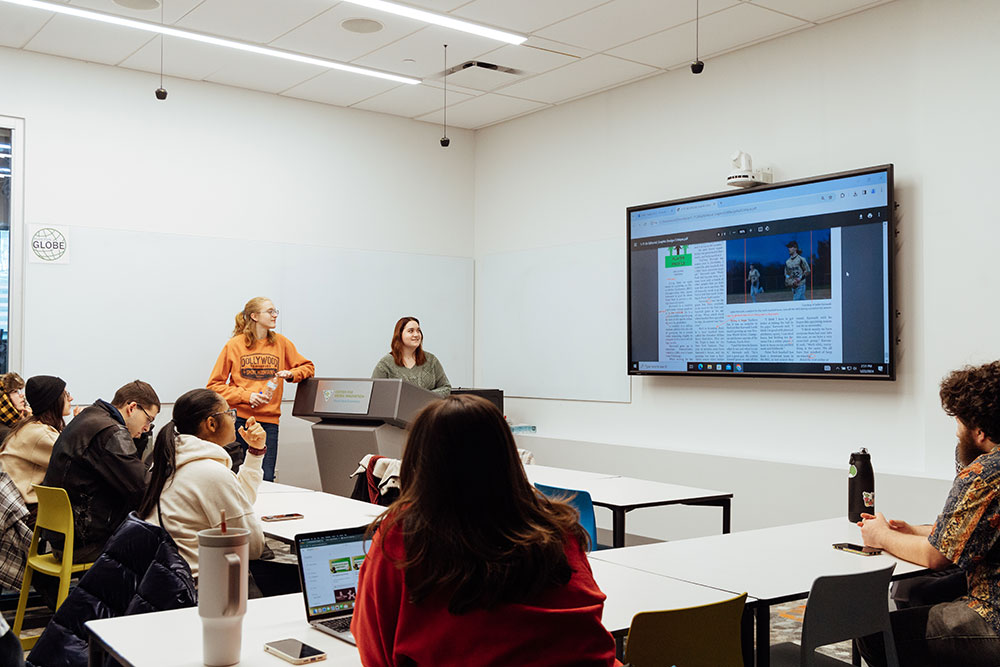 Two students stand at the front of the classroom while others look at the screen.