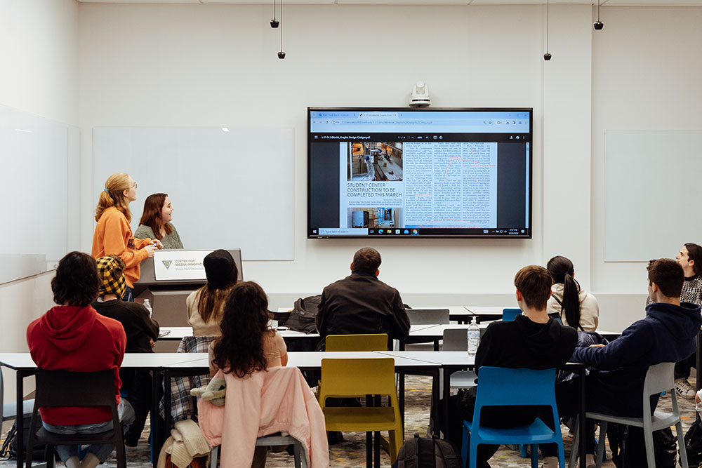Students sit at desks facing a screen on which newspaper layout is shown.