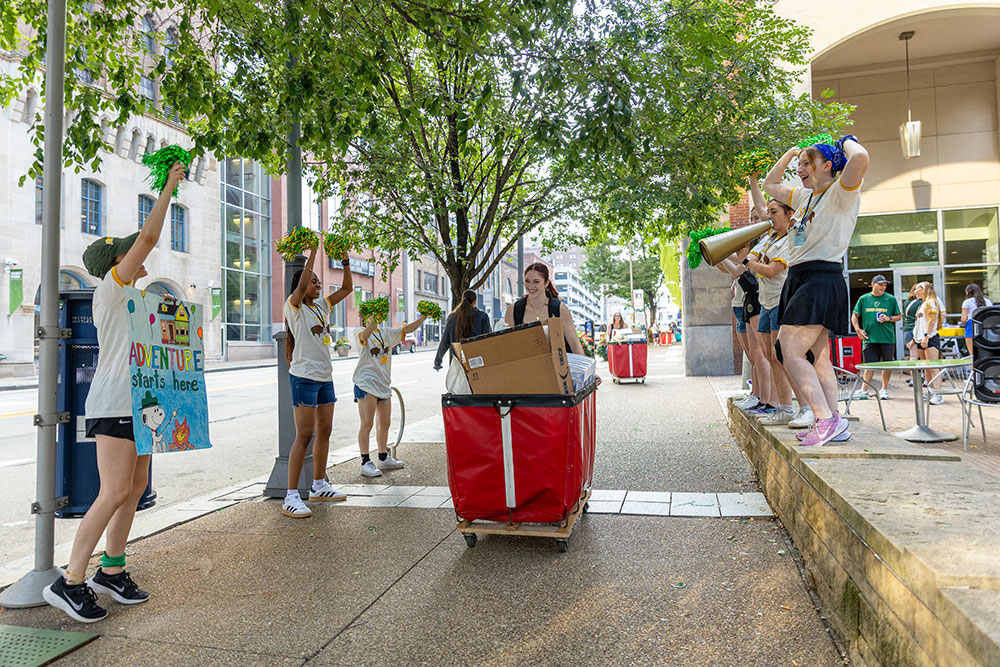 A new student pushes a red bin of her items while older students stand around and cheer.