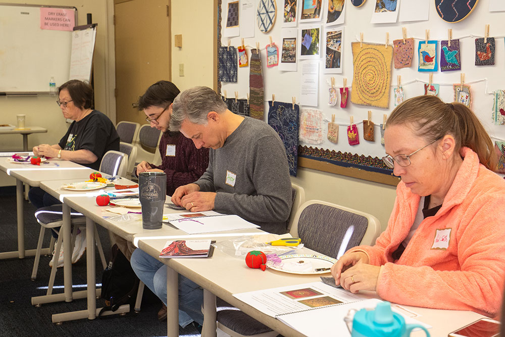 Four people sit at tables and work on embroidery.