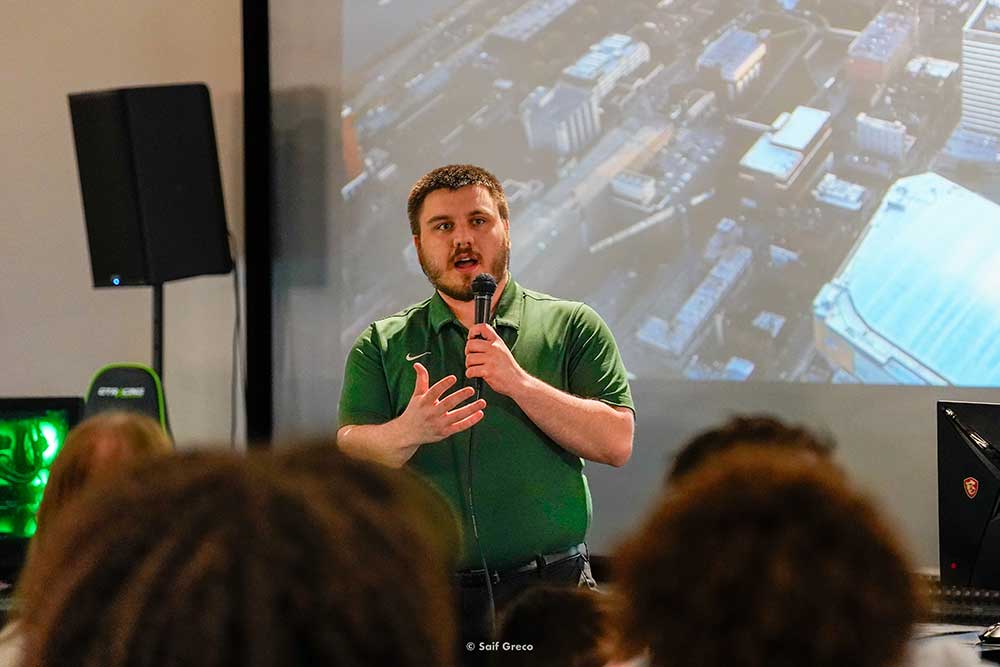 Caleb Knox speaks into a microphone in front of a large screen.