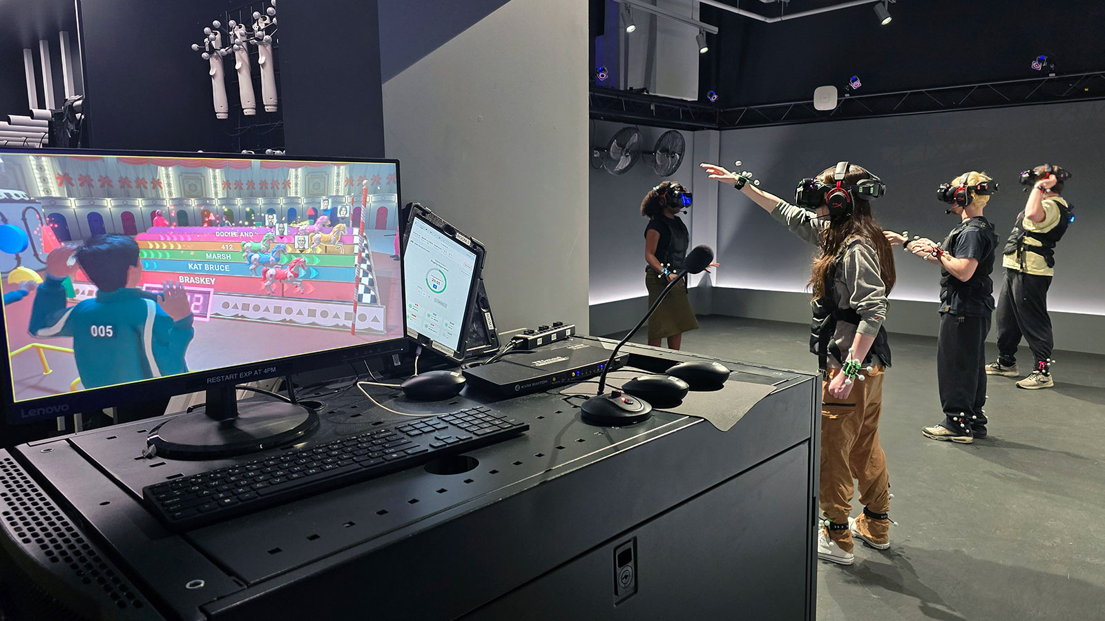 Students stand in a room wearing VR headsets while a game displays on a nearby computer.