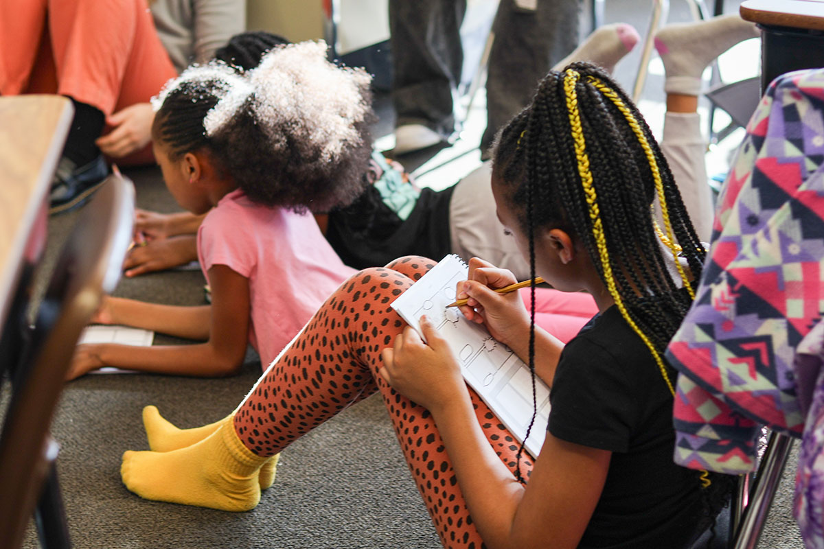 A girl sitting on the floor writes on a worksheet.