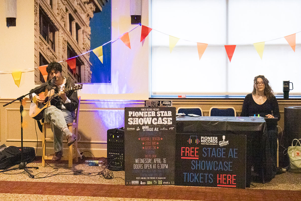 A student plays a guitar next to a table advertising the Pioneer Showcase.