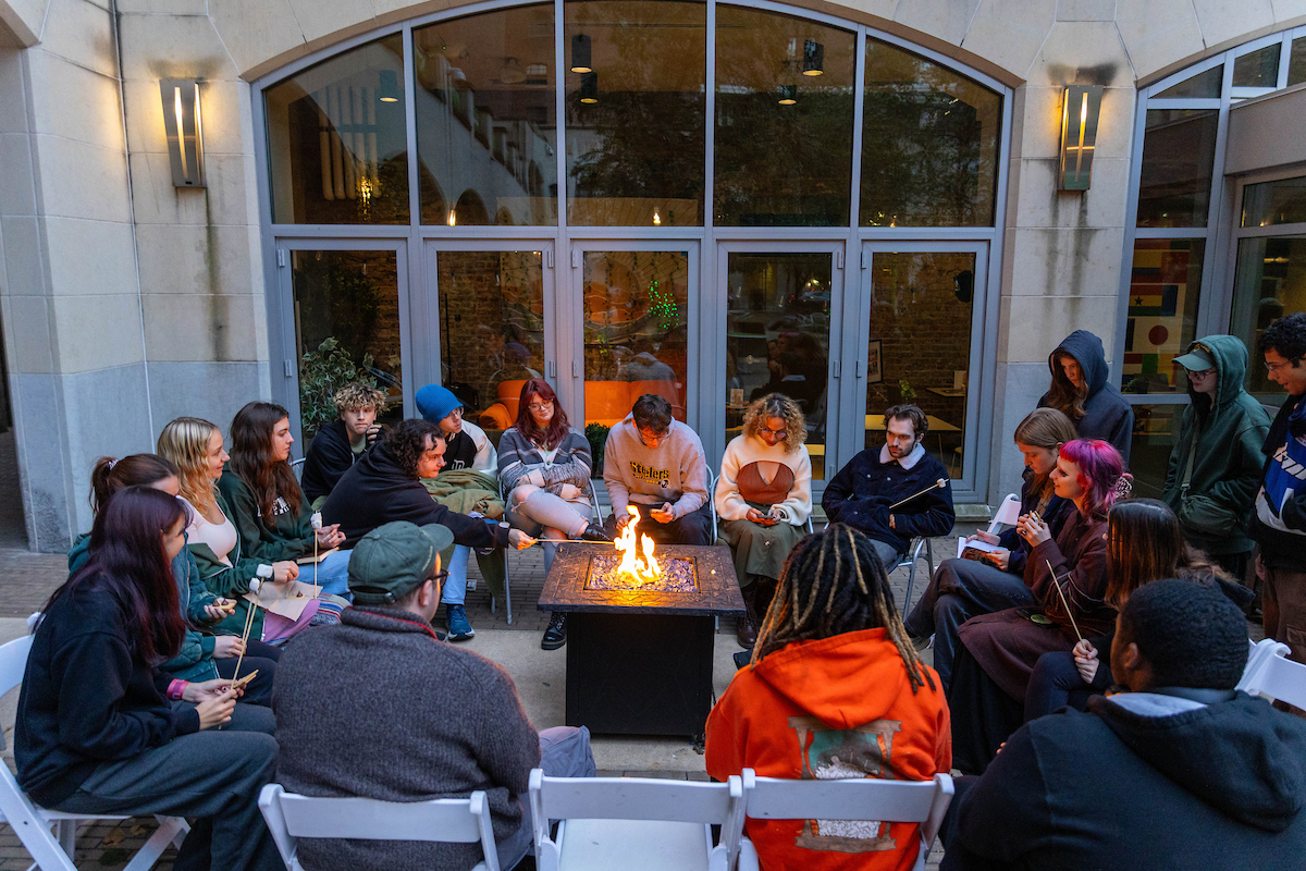 Students sit around a firepit while one reads from his phone. 