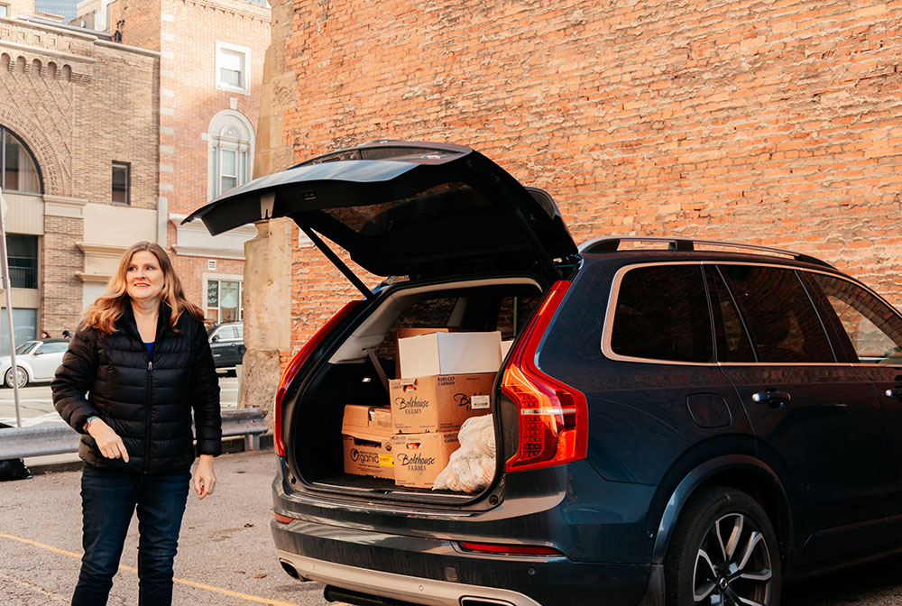 A woman stands by her car, in the open trunk are boxes of produce. 
