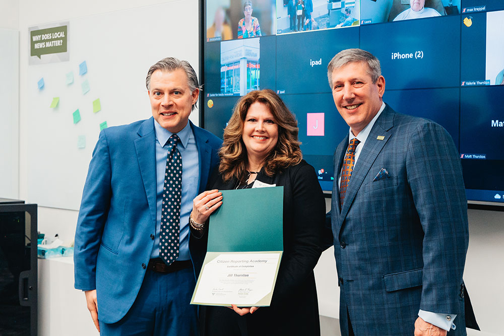 A woman stands between two men, holding her graduation certificate up.