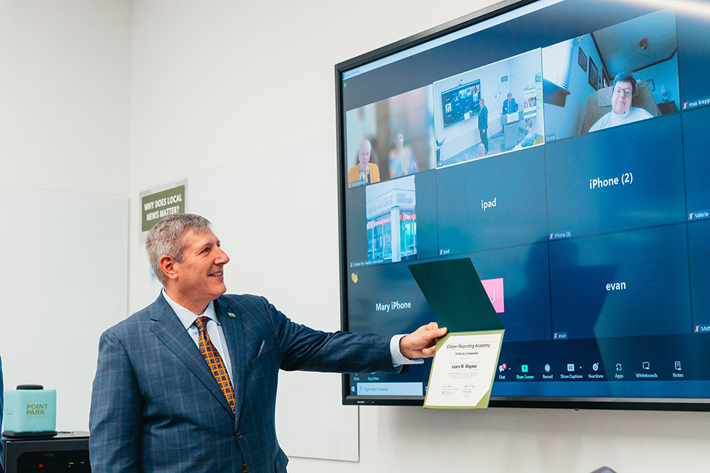 Chris Brussalis stands in front of a screen with people on Zoom.