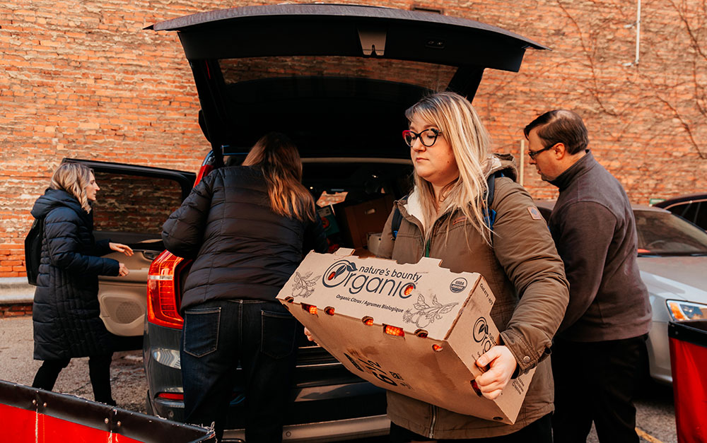 A woman carries a box of food to a red rolling bin.