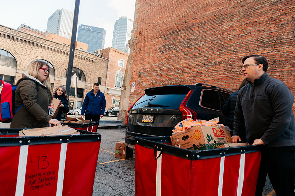 Two people stand in a parking lot with hands on a red bin full of food.