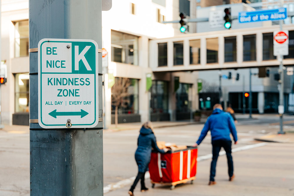 A pole with a "Be Kind" sign is in foreground, in background, two people push a red bin of food across the street. 