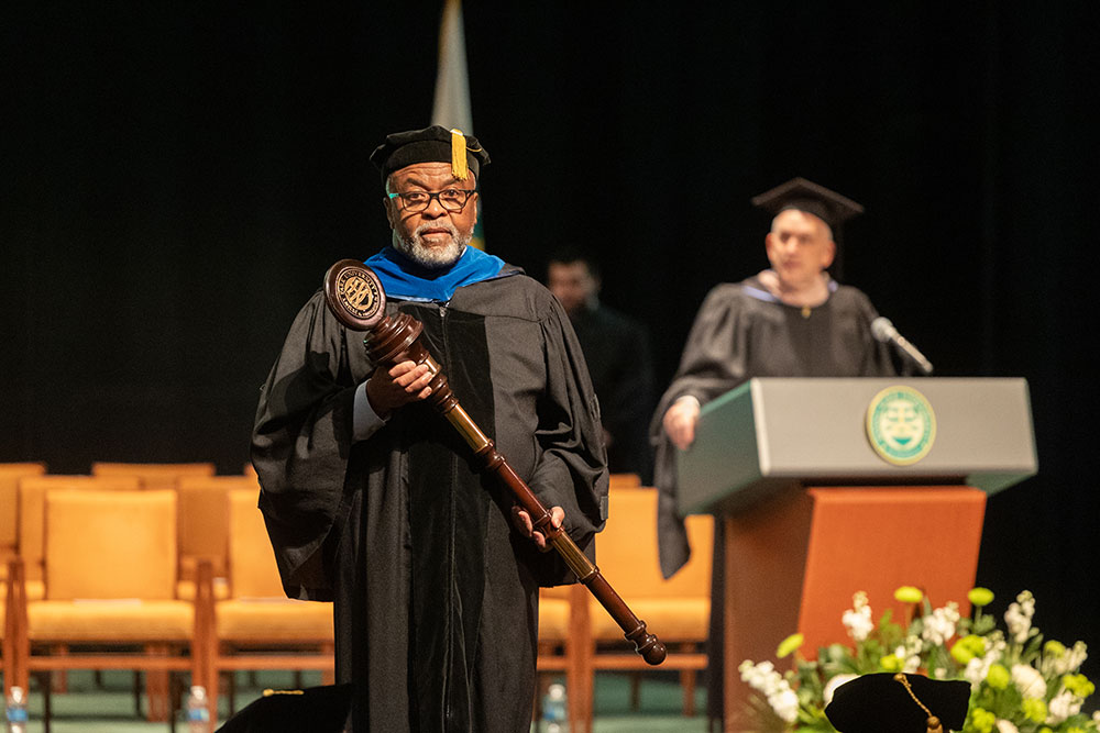 A man in academic regalia holds the university mace on the stage.