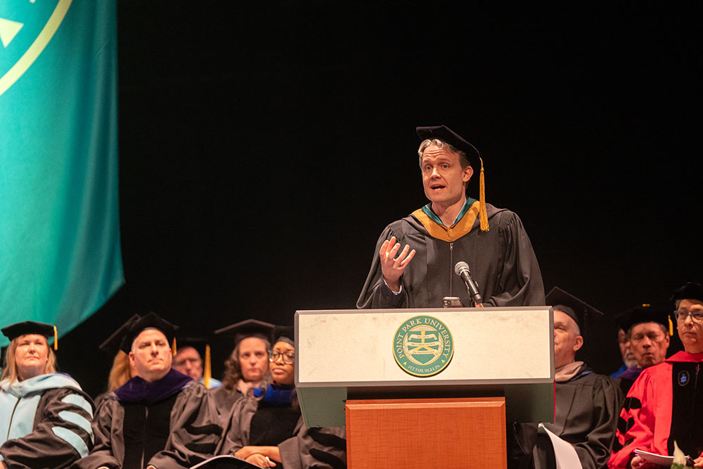 A man in academic regalia speaks from a podium.