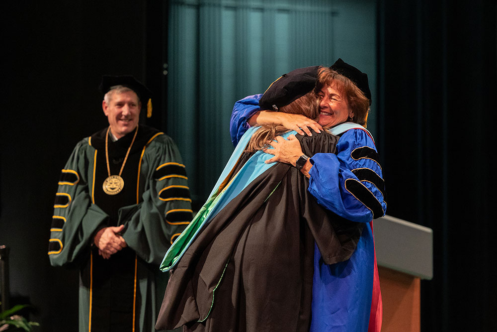 A woman in academic regalia embraces another on stage.