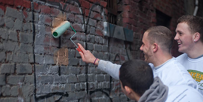 Three male Point Park students use rollers to paint over unsightly graffiti on a brick wall in Downtown Pittsburgh for Pioneer Community Day 2013. | Photo by Christopher Rolinson
