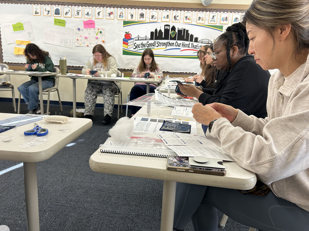 Staff and students handstitch fabric in an Academic Hall classroom.