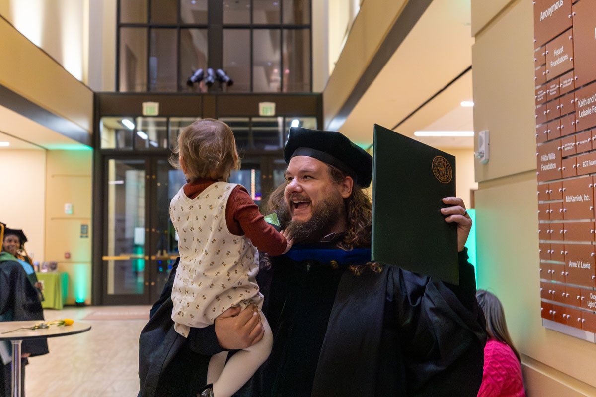 A graduate holds a toddler in one hand and his diploma in the other.