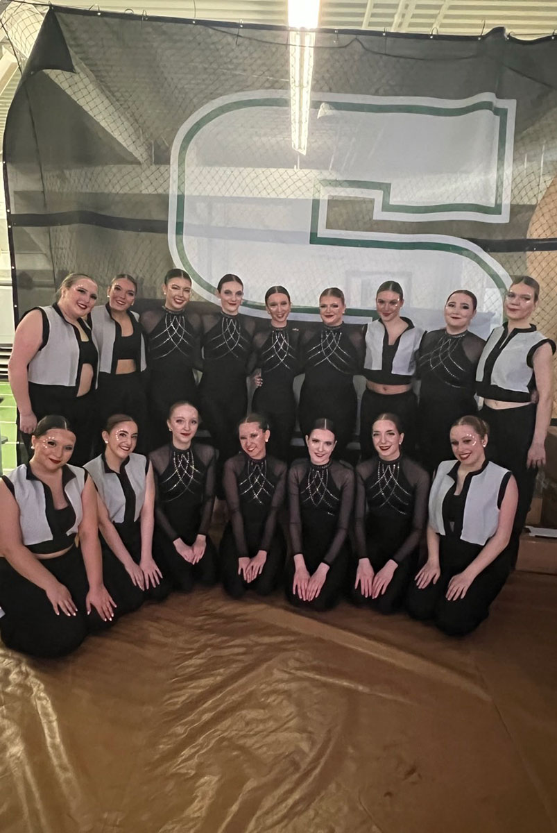 The team poses before a large banner with an S on it.