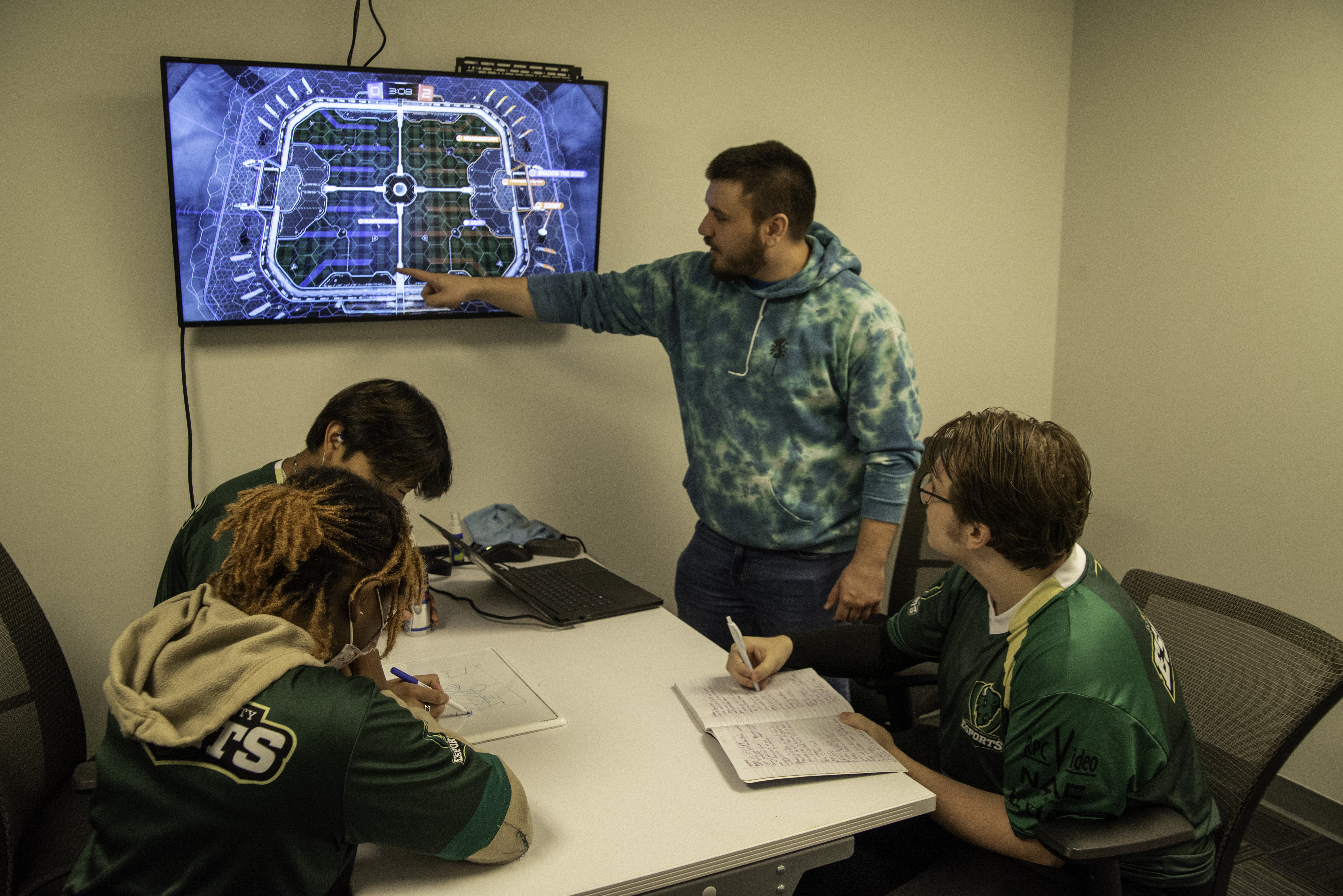 Students sit around a table while a coach points something out on a screen.