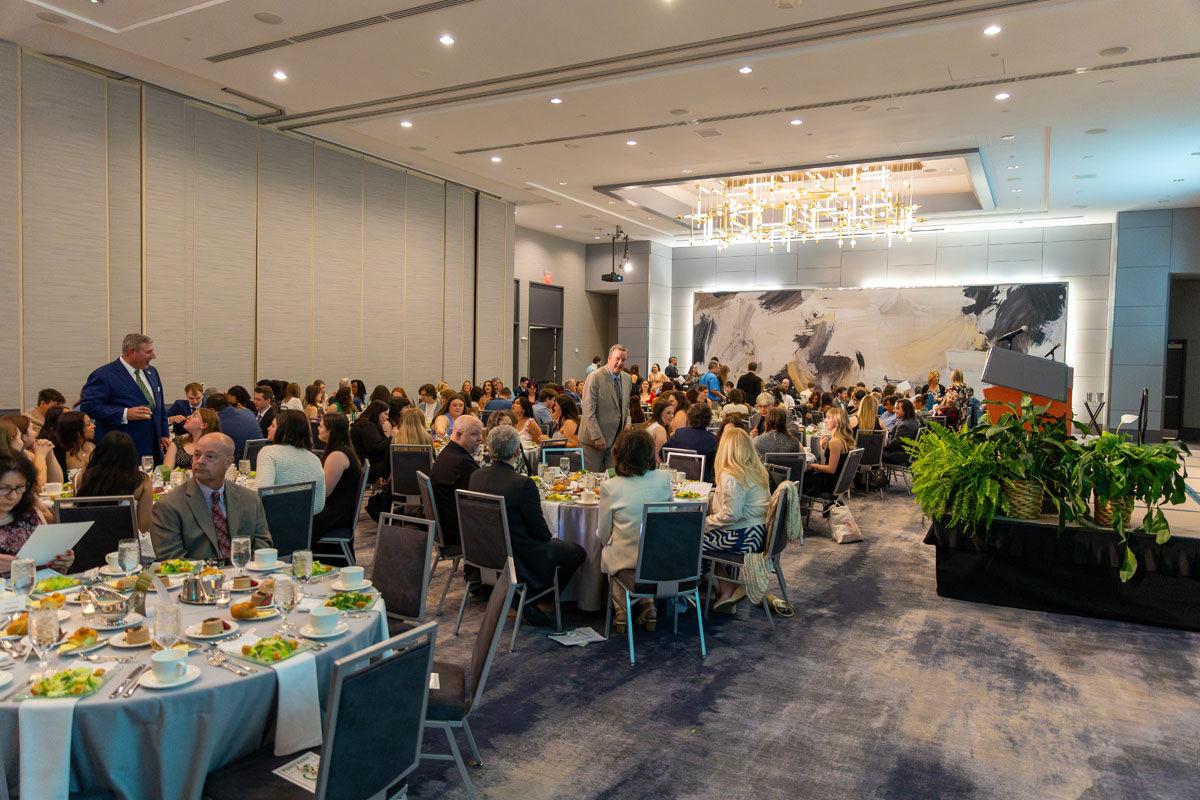 A wide photo of the banquet room filled with tables and people sitting at them. 