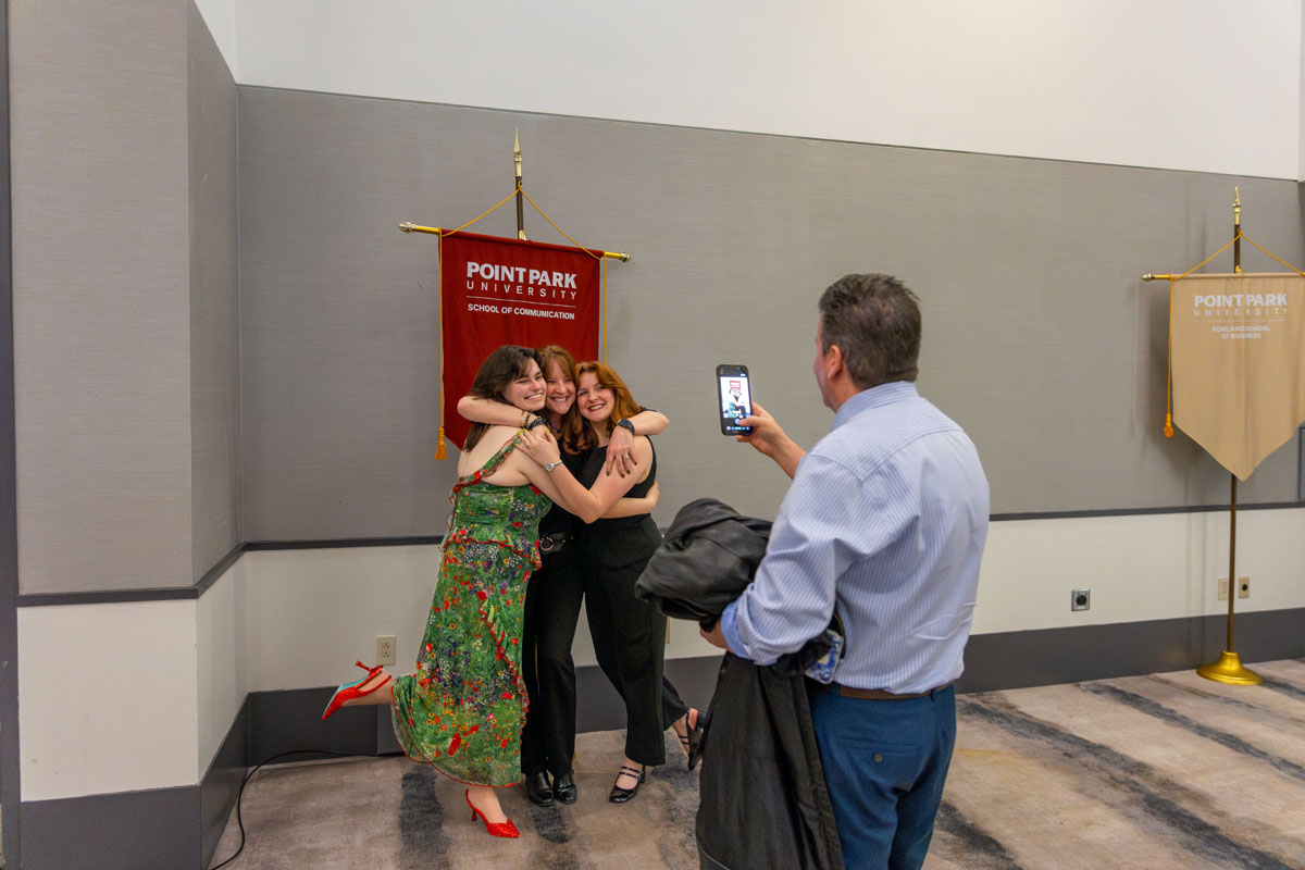 Two students pose for a photo while a man takes their picture on a phone.